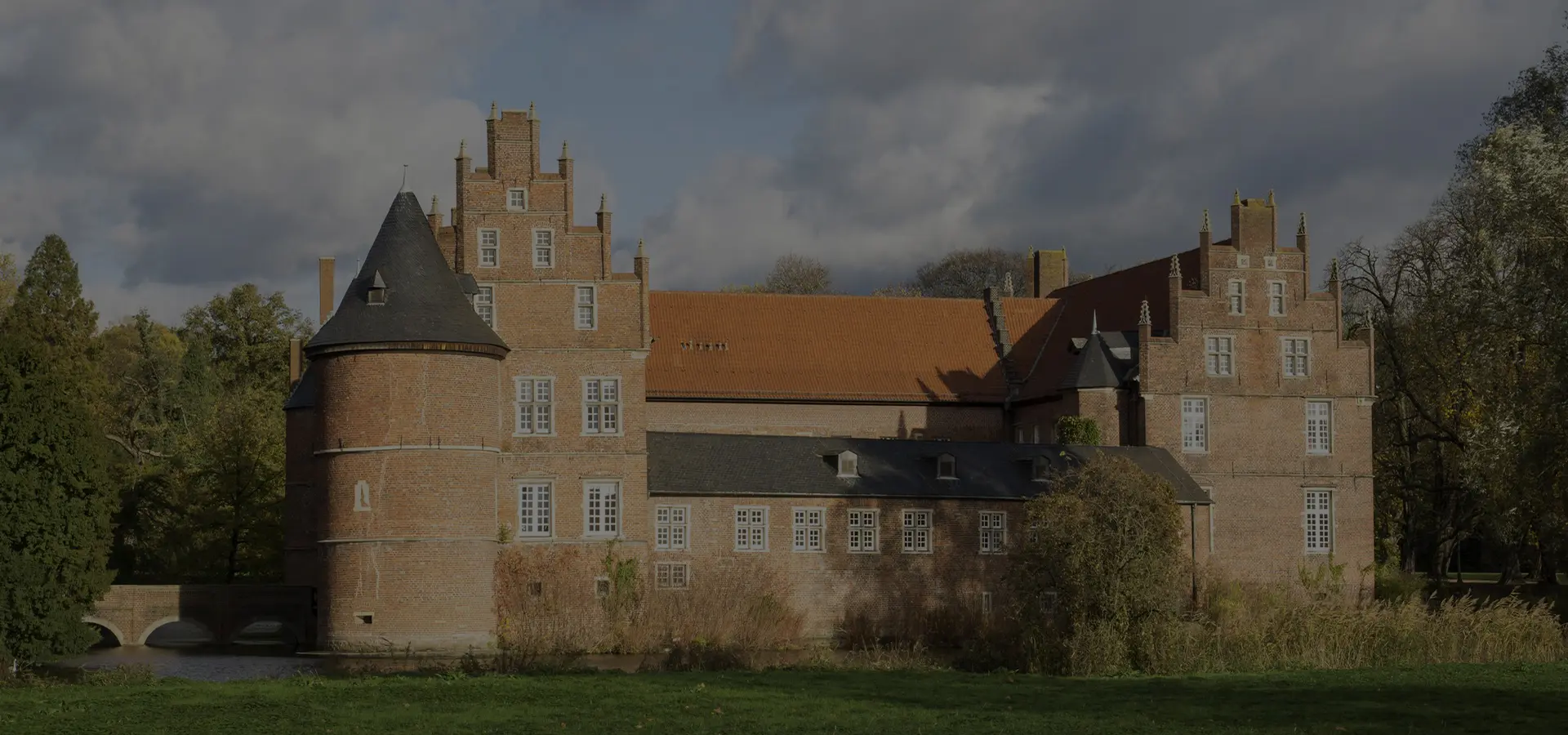 Historisches Schloss mit Ziegelfassade und Türmen, umgeben von Bäumen und Wasser, unter einem bewölkten Himmel.