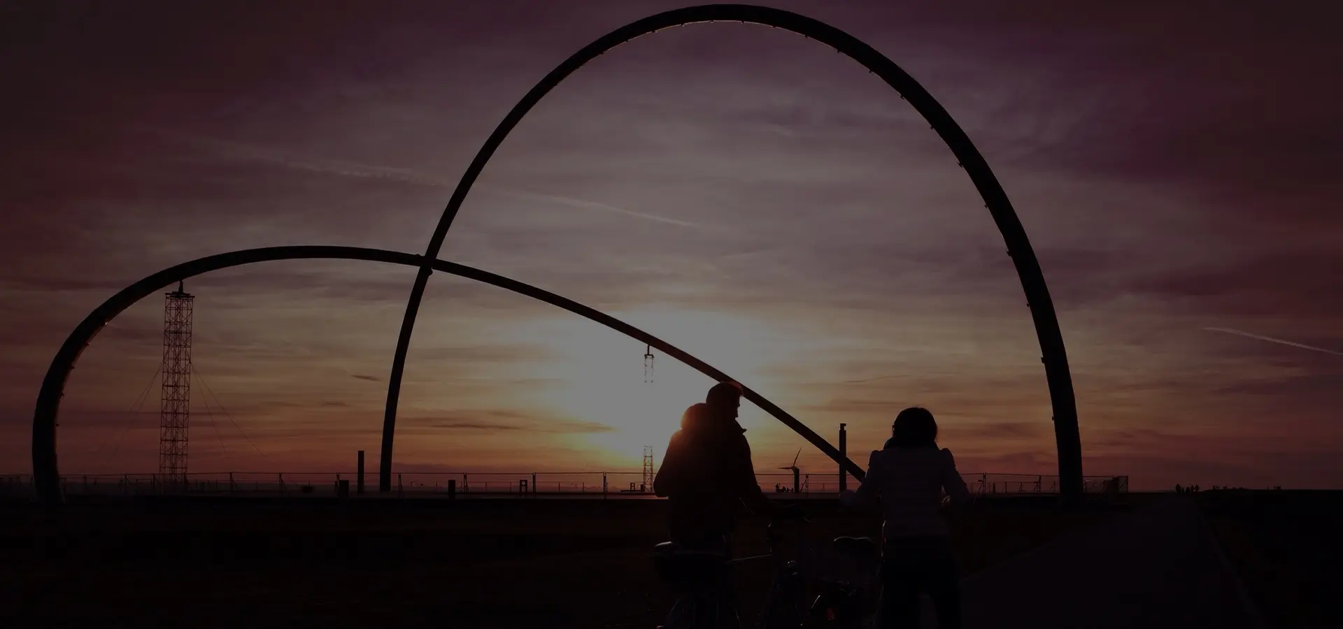 Silhouette von zwei Personen mit Fahrrädern vor einem großen, geschwungenen Metallbogen bei Sonnenuntergang.