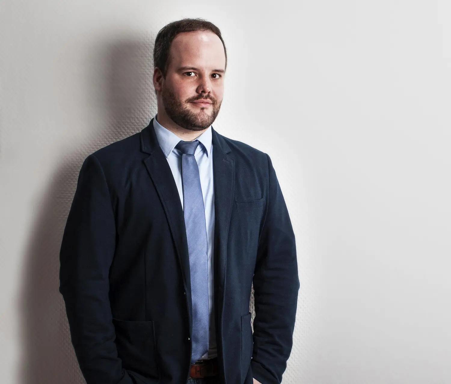 Mann in einem dunklen Blazer und hellblauer Krawatte steht vor einer neutralen Wand.